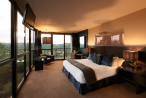 The King Room Mountain View hotel room with a king sized bed, side tables and lamps that have been turned on. A leather side chair in the corner, with many windows and a tv mounted on the wall.