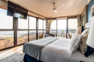 The Junior Suite Mountain View hotel room with large windows with a view of nature from the king sized bed in the centre of the room. There are pillows on the bed and a tv hanging from the celling