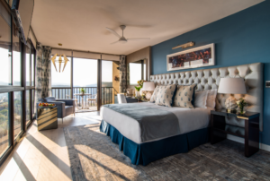 A hotel room with a Mountain View. The hotel room has a king sized bed with a silver head board and blue wall behind. The room has many large windows and a sliding door leading to a balcony with a patio set.