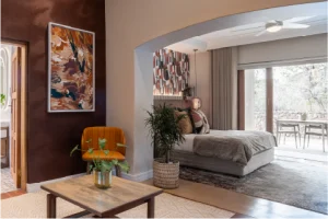 A cozy bedroom with warm earth tones, featuring a plush bed, abstract artwork, and potted plants. A sliding door opens to a bright patio.