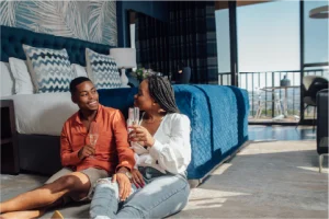 A couple sits on a bedroom floor, smiling at each other while toasting with champagne. The room is bright, featuring a large bed and open balcony view.