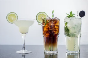 Three cocktails on a reflective surface: a margarita with a lime slice, a dark iced drink with a straw, and a mojito with mint and lime. Refreshing and inviting.