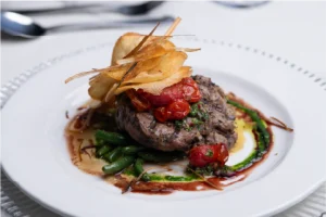 A gourmet dish featuring steak topped with cherry tomatoes and crispy chips, on a plate with green beans and a drizzle of sauce. Elegant and appetizing.