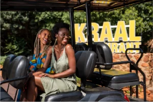 Two women smile joyfully on a vehicle against a backdrop of greenery and a yellow restaurant sign. They exude happiness and warmth in the sunlit setting.
