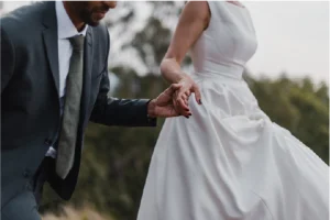 A couple in formal attire walks hand in hand outdoors. The woman wears a flowing white dress; the man is in a suit. The setting is serene with blurred trees.