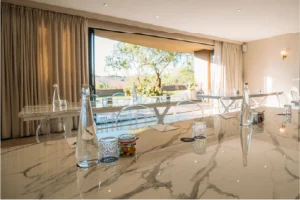 Spacious room with marble tables set with water bottles and glasses. Open patio doors reveal a sunny garden view, creating a serene atmosphere.