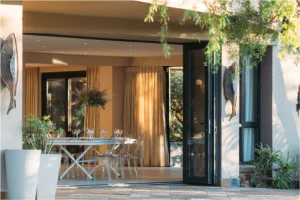 Modern dining area with glass table and chairs, adorned with wine glasses, opens to a patio. Warm lighting and decor create a welcoming ambiance.
