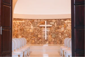 An elegantly lit chapel with white chairs facing a stone wall adorned with a large white cross and two menorahs. The mood is serene and reverent.