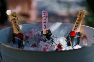 A bucket filled with ice holds three bottles of Moët champagne. Pink and gold labels are visible, surrounded by ice cubes, creating a celebratory, elegant ambiance.