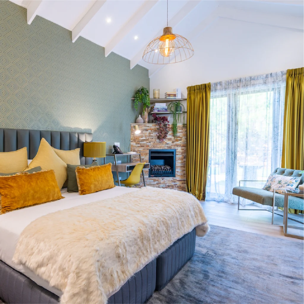 A cozy bedroom with soft lighting features a large bed with a fluffy throw and mustard pillows, a modern fireplace, and floor-to-ceiling curtains.