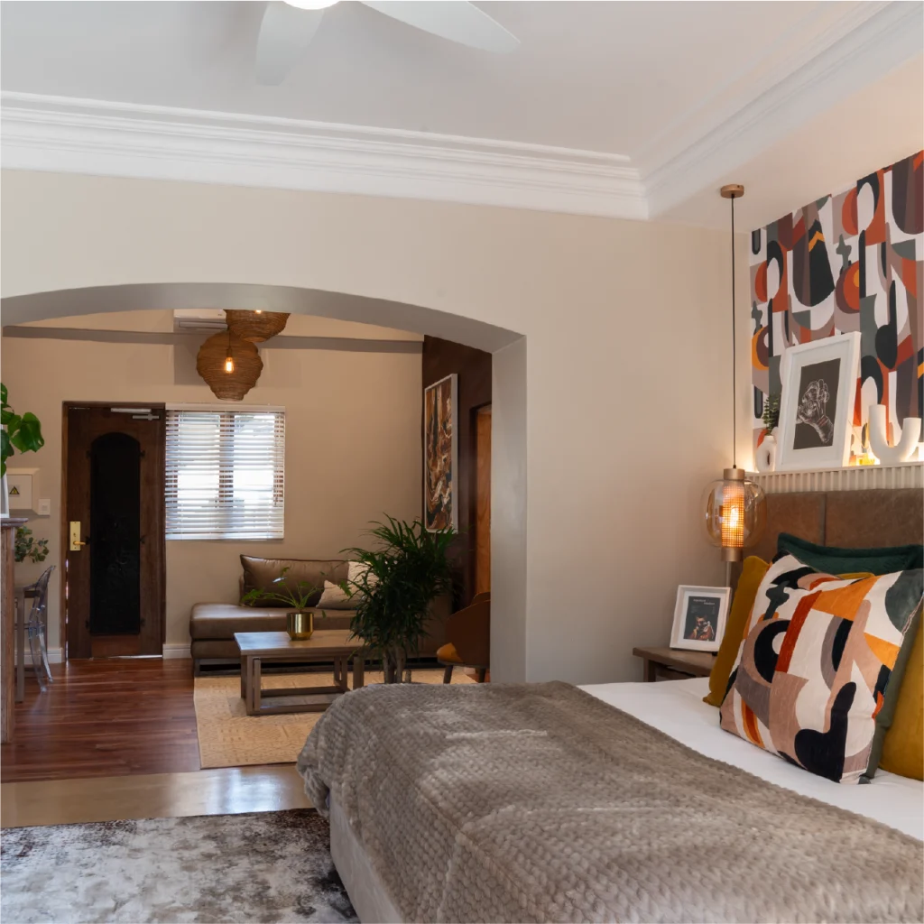 A cozy bedroom with modern decor; features a large bed with colorful pillows, abstract art on walls, and a view into a warmly lit sitting area with potted plants.